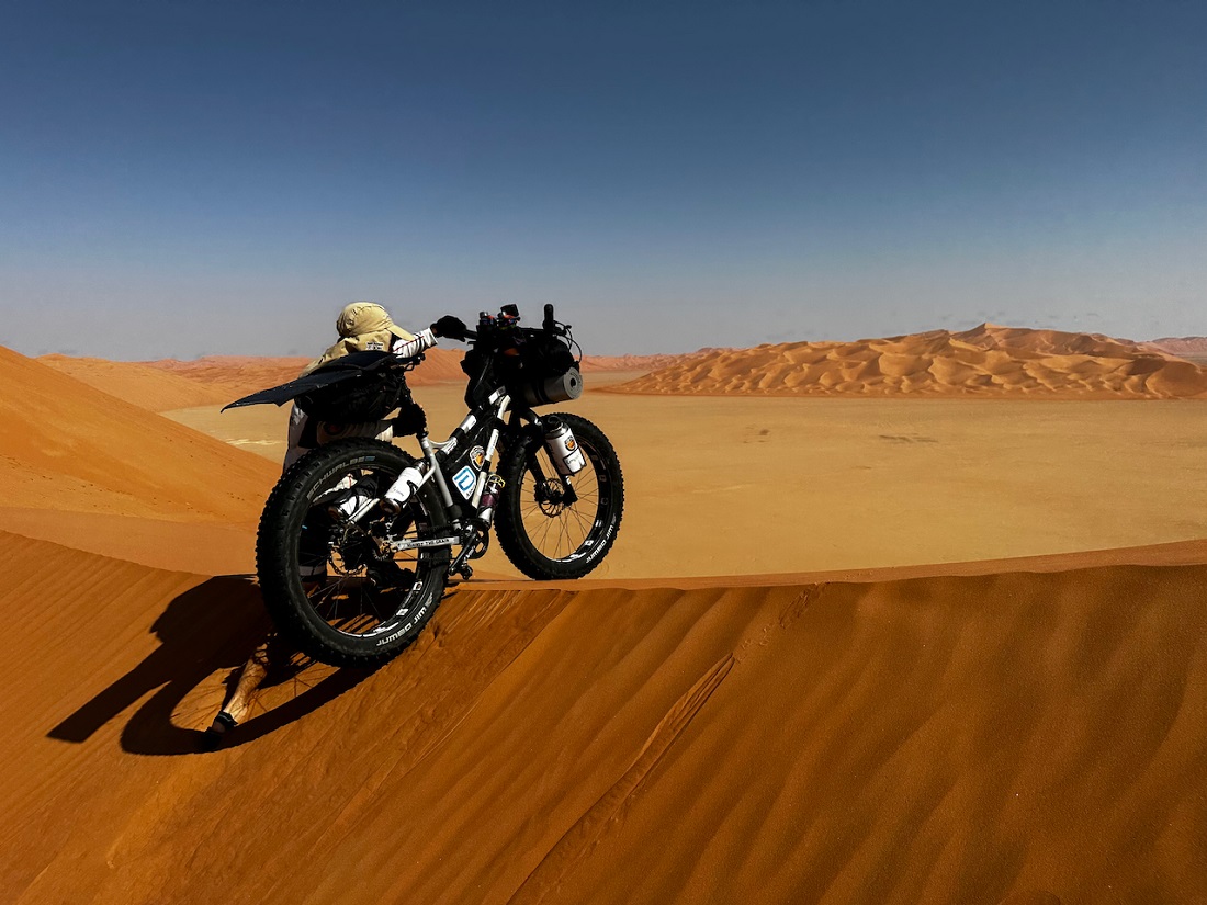 a man pushes a heavily laden bike up and over a high sand dune