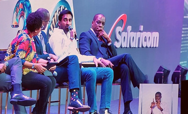 four men are seen on a presentation stage, with one of them talking into a mic.