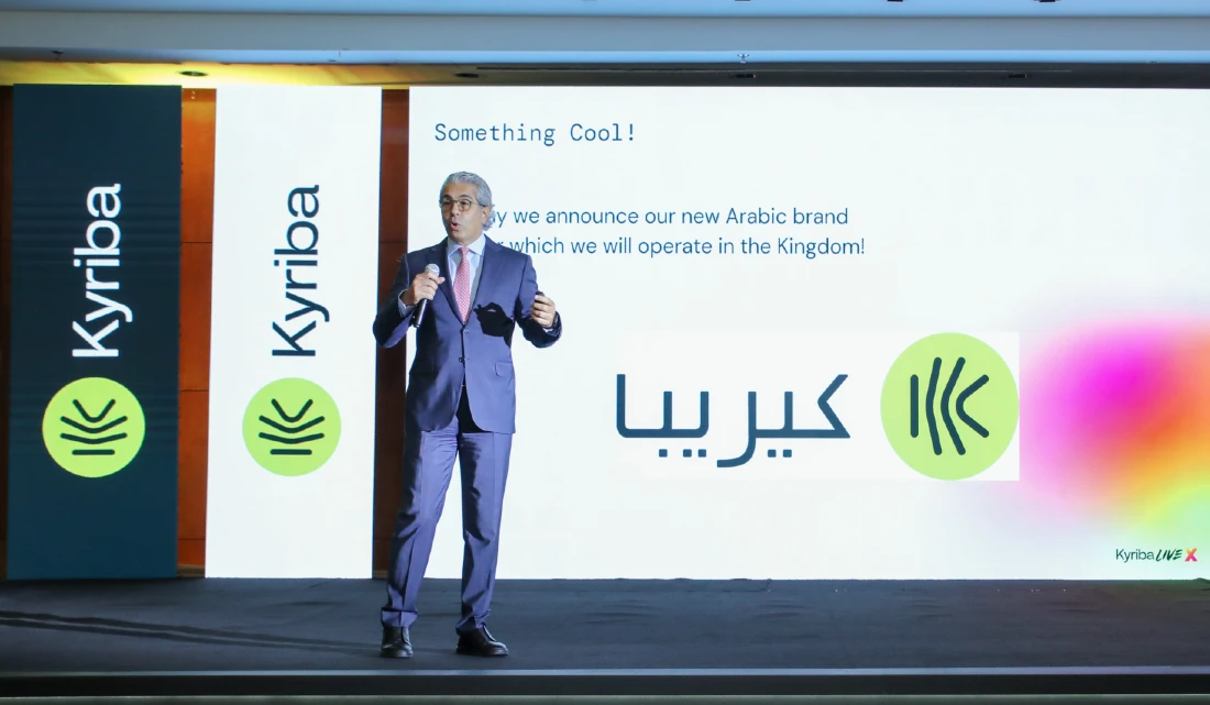 a man is seen on stage talking into a mic in front of a presentation screen with english and arabic text upon it