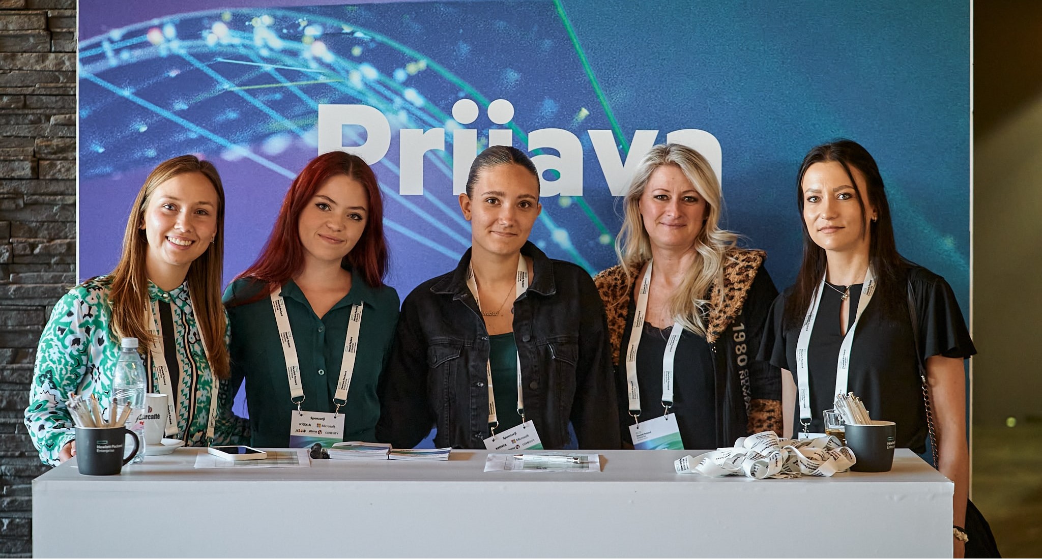 Five women are seen in a group photograph with each of them wearing a lanyard. They are pictured behind a welcome desk.